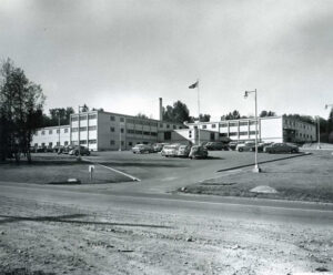Low-raise institutional building with vintage 1960s cars parked in front