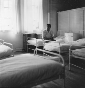 man sitting along on bed in large institutional dorm