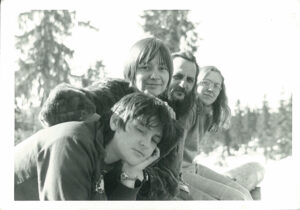 3 young women and a man with long hair and a beard all sitting together with the woods behind.
